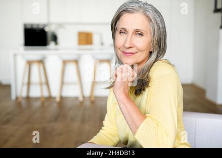 Elegante e affascinante donna di mezza età con capelli grigi seduti sul comodo divano e guarda la macchina fotografica, anziana signora sorridente, mettere il suo mento a portata di mano, trascorrere il tempo libero a casa. Concetto di benessere Foto Stock