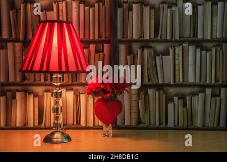 Lampada da tavolo con ombra rossa e un mazzo di rose rosse con cuori rossi davanti ad un libro Foto Stock