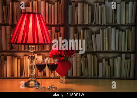 Lampada da tavolo con paralume rosso, due bicchieri pieni di vino rosso e un mazzo di rose rosse con cuori rossi davanti ad un libro Foto Stock