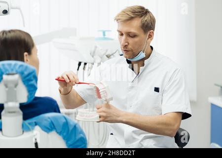 Dentista che mostra la corretta igiene dentale utilizzando mock-up di scheletro di denti. Medico stomatologo che spiega la corretta igiene dentale per il paziente che tiene un campione di mandibola umana con spazzolino da denti. Foto Stock