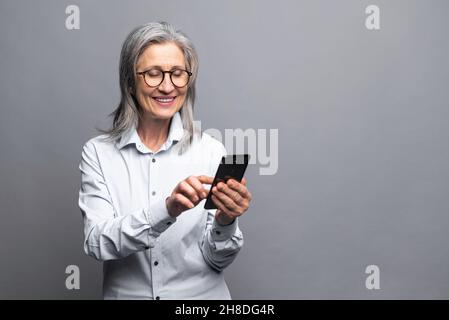 Allegro positivo felice anziano donna d'affari grigio-pelata usando smartphone in piedi isolato su grigio, donna matura in abbigliamento formale tocco touchscreen, scorrimento news feed, messaggi, SMS Foto Stock