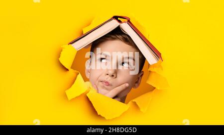 Ragazzo caucasico pensivo con libro sulla testa che pensa, peping attraverso il foro in carta arancione strappata, disegno del banner Foto Stock