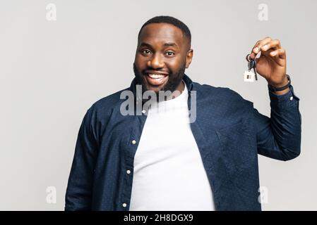 Ragazzo felice che tiene le chiavi con portachiavi in forma di piccola casa isolato su sfondo grigio. Sorridente uomo afroamericano tiene le chiavi dalla nuova proprietà, compratore felice della propria proprietà. Un ragazzo si è riposizionato Foto Stock