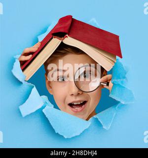 Ragazzo carino e intelligente con lente d'ingrandimento che si nasconde sotto il libro, guardando attraverso il foro in carta blu strappata Foto Stock