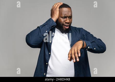 Scoraggiato ragazzo afroamericano lettura notifica sullo schermo di smart watch, in piedi isolato su grigio, deluso uomo nero in casual look alla smart band sul suo polso, in ritardo per la riunione Foto Stock