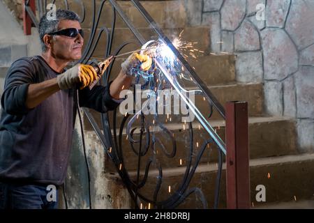 Uomo che indossa occhiali protettivi salda metallo con saldatrice in una casa privata. Le scintille provenienti dalla saldatura volano sui lati. Foto Stock