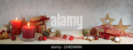 Decorazioni natalizie in formato panoramico con candele rosse, baubles, nastro, stelle in legno e regali avvolti in natura su un tavolo, parete grigio chiaro con co Foto Stock