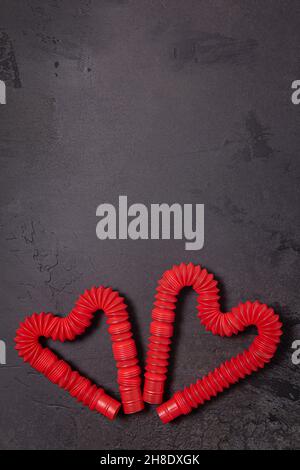 Steso rosso pop tubo bambini giocattoli piegati a forma di due cuori. Giocattoli antistress in plastica su sfondo di pietra nero testurizzato. Disposizione piatta. Copia Foto Stock