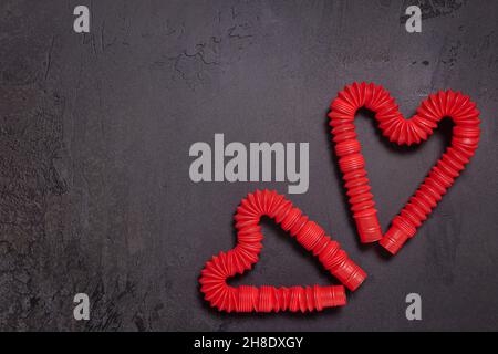 Due stese rosso pop tubo per bambini giocattoli piegati a forma di cuori. Giocattoli antistress in plastica su sfondo di pietra nero testurizzato. Disposizione piatta. Copia Foto Stock
