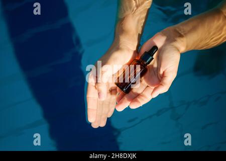 Donna mano che tiene vuoto siero organico Skincare prodotto vetro ambrato bottiglia per l'uso in un giorno con fondo acqua piscina blu chiaro semplice. Foto Stock