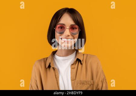 Ritratto di giovane donna positiva in occhiali da sole eleganti colorati, sorridendo alla macchina fotografica su sfondo giallo studio Foto Stock