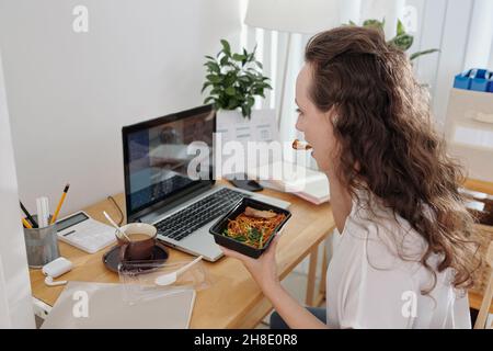 Giovane donna d'affari seduta alla scrivania d'ufficio, pranzo e ascolto di un collega durante la conferenza online Foto Stock
