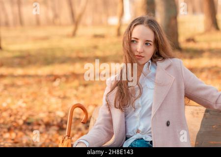 Una ragazza da sola siede con un ombrello nelle sue mani su una panchina in un parco autunnale Foto Stock