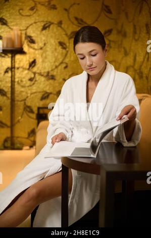Fidata attraente e rilassata giovane donna che riposa in una zona lounge di una clinica wellness spa, indossando accappatoio, rivista di lettura e preparandosi per r Foto Stock