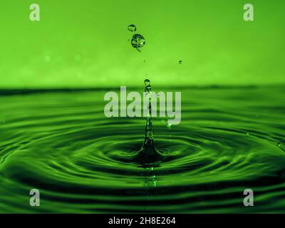 Una goccia verde gocciola nell'acqua e crea spruzzi di varie forme, a causa degli spruzzi, le onde sono create sull'acqua, il concetto di un liquido Foto Stock