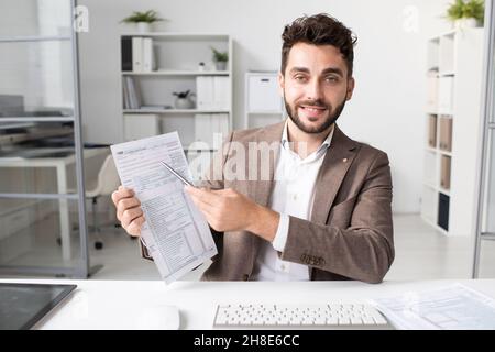 Ritratto di ragioniere sorridente barbuto in giacca seduta alla scrivania in ufficio moderno e indicando la forma di imposta Foto Stock