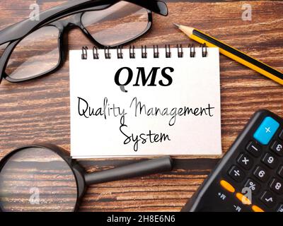 Occhiali, calcolatrice, matita e taccuino con testo QMS Quality Management System su un tavolo in legno Foto Stock