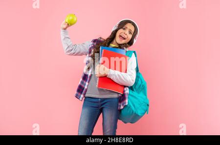 il bambino scioccato ascolta la musica in cuffie portare zaino con libro degli esercizi e mela, pranzo. Foto Stock