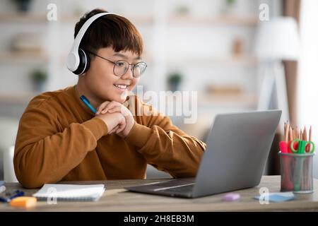 Cheerful ragazzo asiatico scuola che ha una lezione online da casa Foto Stock