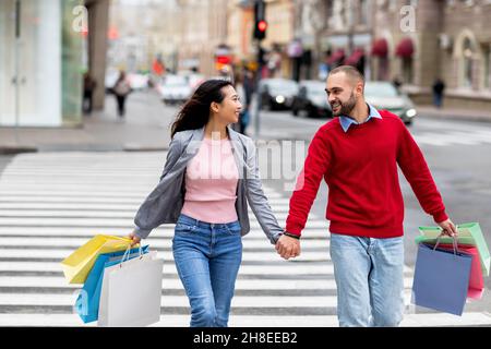 Ritratto di giovane coppia internazionale con borse regalo colorate che attraversano la strada, tenendo le mani all'aperto Foto Stock