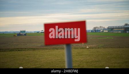 Focus dietro il luminoso rosso airfield attivo segno pista sugli edifici a campi d'aria e oltre la pista di erba, Wiltshire UK Foto Stock