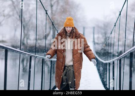 Una donna cammina sul fiume su un ponte sospeso in inverno. Giovane ragazza in abiti caldi si trova su un ponte di legno in una fredda giornata innevata. Foto Stock