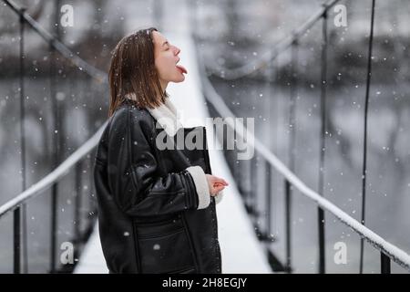 Una donna cammina e cattura fiocchi di neve con la lingua sopra il fiume su un ponte sospeso in giorno d'inverno. Giovane ragazza in abiti caldi si alza su un legnino Foto Stock