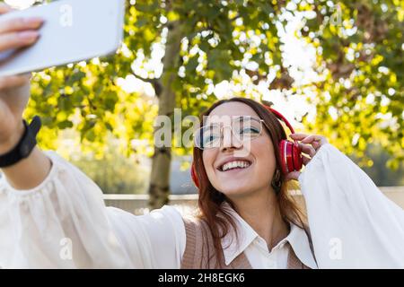 Donna d'affari rossa positiva con occhiali che prendono selfie via smartphone mentre ascoltano musica e sorridono nel parco. Musica e natura concetto Foto Stock