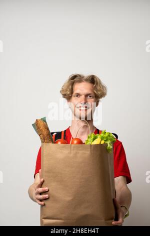 Ritratto di felice uomo di consegna in rosso uniforme sorridente mentre trasportano il sacchetto di carta con prodotti alimentari isolati su sfondo bianco. Foto Stock