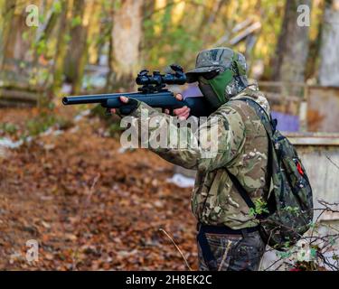 Airsoft Sniper prendendo la mira attraverso l'ambito di un bersaglio sconosciuto nella foresta, il suo fucile è nero e indossa mimetizzazione, protezione occhi & viso Foto Stock