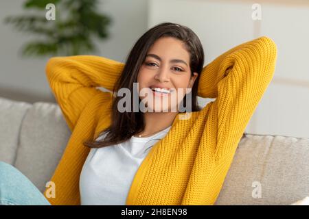 Riposatevi e rilassatevi a casa vostra. Una donna musulmana felice gettò le mani dietro la testa e sorrise alla macchina fotografica, seduto sul divano Foto Stock