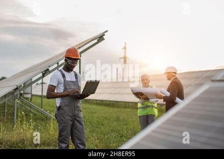 Tecnico afroamericano in uniforme in piedi sul campo con pannelli solari e digitando su laptop, mentre donna musulmana e uomo indiano che lavorano con i progetti dietro. Concetto di energia alternativa. Foto Stock