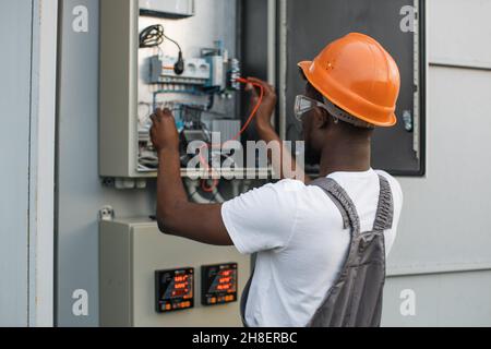 African american riparatore in casco arancione e occhiali di sicurezza controllo cavi in trasformatore di potenza tra stazione solare. Tecnico competente che controlla il processo di produzione di energia verde. Foto Stock