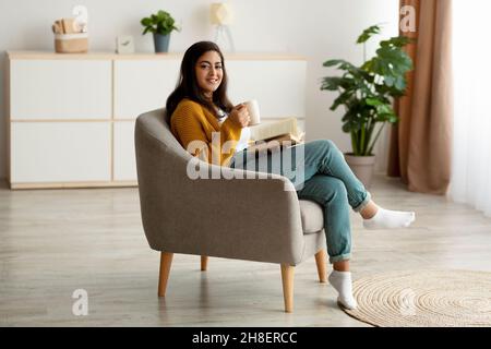 Passatempo fine settimana. Felice donna araba leggere libro, bere caffè e sorridere alla macchina fotografica, seduto in comoda poltrona Foto Stock