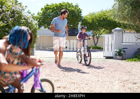 Padre e figlio corrono e pedalano in un vialetto soleggiato Foto Stock