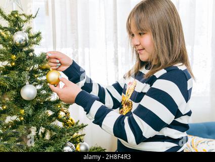Ragazza bionda sorridente che mette una sfera di natale sull'albero. Decorare la casa per celebrare il Natale Foto Stock