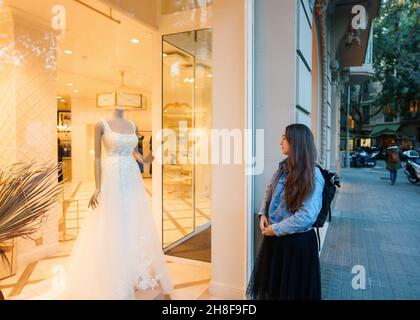 Giovane donna in piedi vicino vetrata del negozio in città e sognando su abito da sposa bianco Foto Stock