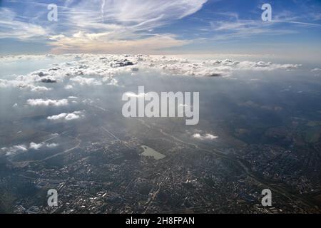 Vista aerea dalla finestra dell'aeroplano ad alta quota di città lontana coperta da strato di smog sottile e nubi distanti. Foto Stock