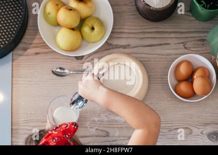 Mani di bambina in fascia rossa cuocere torta di mele in cucina. Il bambino mette lo zucchero nel misurino. I bambini aiutano sui lavori domestici. Cibo da cucina per bambini Foto Stock