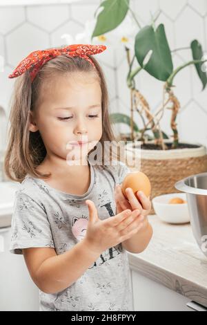 Piccola ragazza dai capelli scuri di 3 anni in banda rossa covata torta di mele in cucina. Il bambino tiene l'uovo nelle mani. I bambini aiutano sui lavori domestici. Bambino Foto Stock