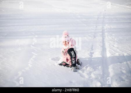 La bambina è seduta nella neve. Il bambino in giacca rosa invernale e cappello con pompom si diverte a piedi nella natura e gode di tempo soleggiato e gioca con la neve Foto Stock