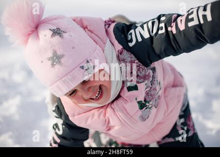 Ritratto del bambino in primo piano. Bambina in giacca rosa invernale e cappello con pompom ha divertimento a piedi nella natura e gode di tempo soleggiato e gioca con Foto Stock