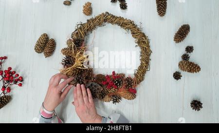 Vista dall'alto delle mani femminili che fanno una corona per natale. Una donna anziana realizza decorazioni per il nuovo anno con le proprie mani Foto Stock