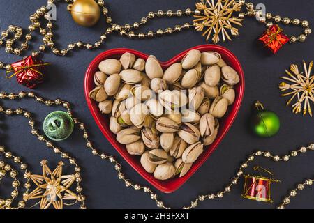 Pistacchi arrostiti salati con guscio rosso a forma di cuore su sfondo nero con decorazioni natalizie, concetto di cibo vegano sano e snack durante le vacanze invernali. Primo piano, messa a fuoco selettiva, spazio di copia Foto Stock