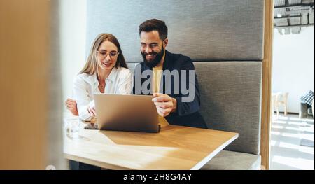 Due professionisti che sorridono a uno schermo di un notebook allegro. Giovani uomini d'affari felici che lavorano insieme in uno spazio di lavoro moderno. Due anni di successo Foto Stock