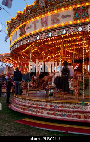 Classic Carousel, Merry Go Round, al Goodwood Revival vintage event 2014, al crepuscolo con luci. Giostra di divertimento con gente in costume Foto Stock