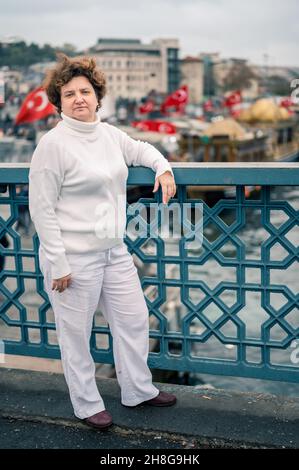 Donna di Ederly in abiti bianchi sul ponte Galata il giorno nuvoloso. Bandiere turche volano sullo sfondo. Una donna di 50 anni, corta, guarda nella macchina fotografica , Foto Stock