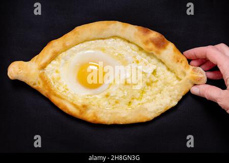 Il piatto nazionale georgiano è il khachapuri adjariano. Mano che tiene aperto formaggio a forma di barca e torta di uova isolato su sfondo nero, primo piano. Nazionale c Foto Stock