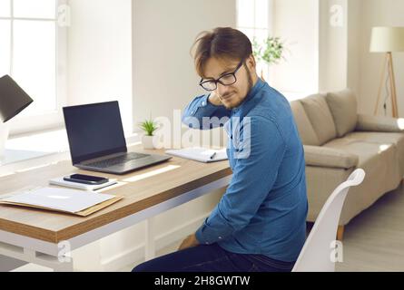 Uomo stanco che ha dolore al collo seduto alla sua scrivania e che lavora sul suo computer portatile Foto Stock