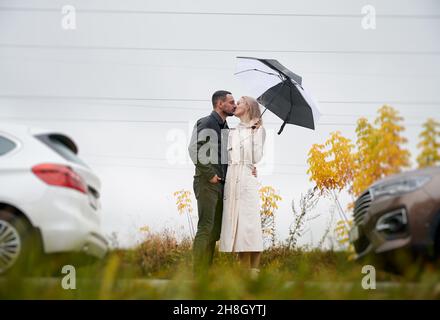 Giovane coppia innamorata baciante teneramente, in piedi sotto ombrello su strada tra due auto su sfondo di erba, cespugli gialli, cielo grigio e cavi elettrici. Romantica atmosfera autunnale. Foto Stock